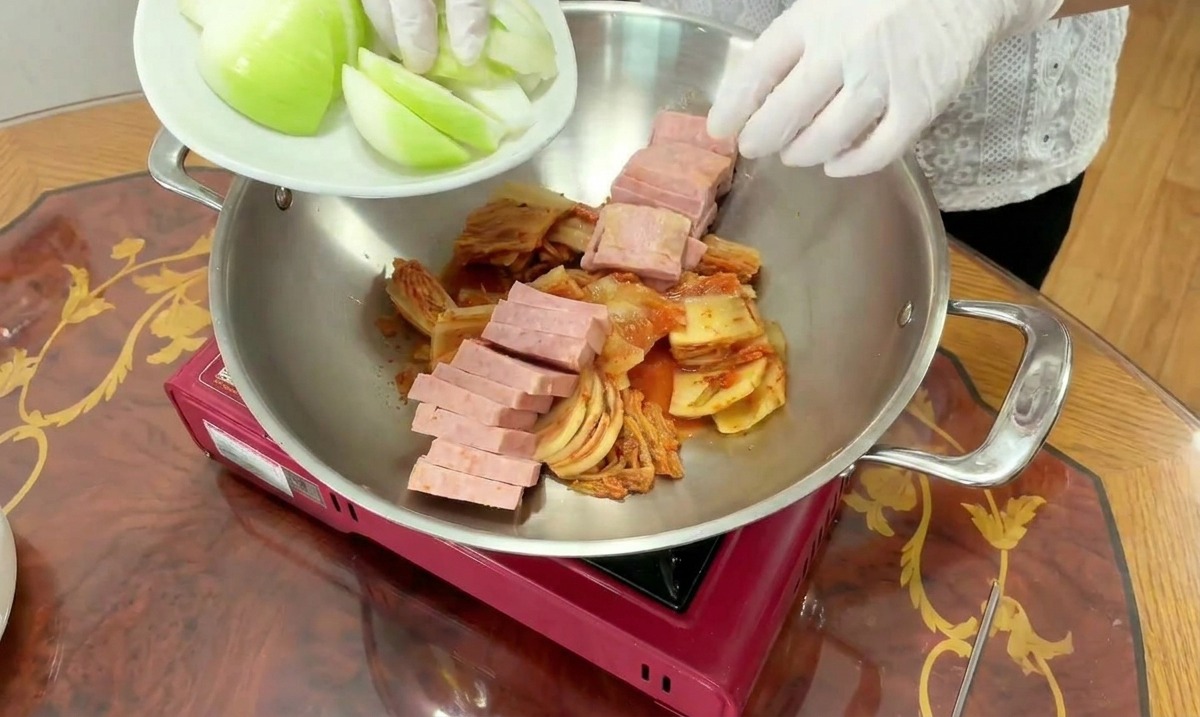 Un cuisinier ajoute des quartiers doignons blancs frais dans une marmite contenant déjà du kimchi et du spam.