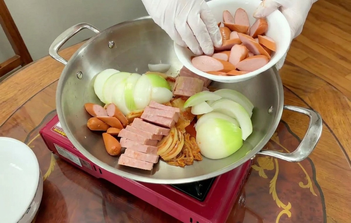 Des saucisses à la peau rouge coupées en rondelles sont versées dun bol blanc dans une marmite avec oignons, jambon et kimchi.