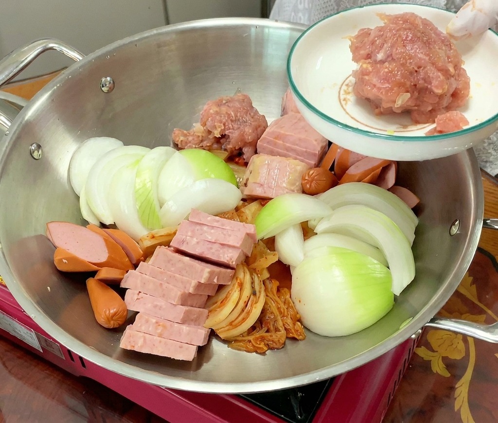 Un monticule de porc haché rose assaisonné est ajouté à la marmite remplie de kimchi, spam, saucisses et oignons.