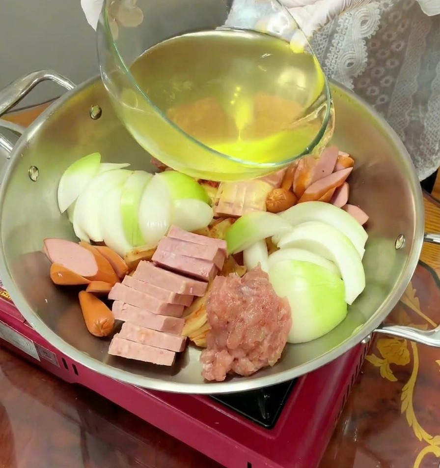 Un liquide jaune clair est versé dun bol en verre dans une marmite en inox remplie dingrédients crus.
