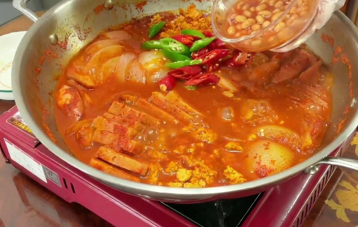 Une main verse des haricots à la tomate dun petit bol en verre dans un ragoût rouge bouillonnant contenant déjà des piments émincés.