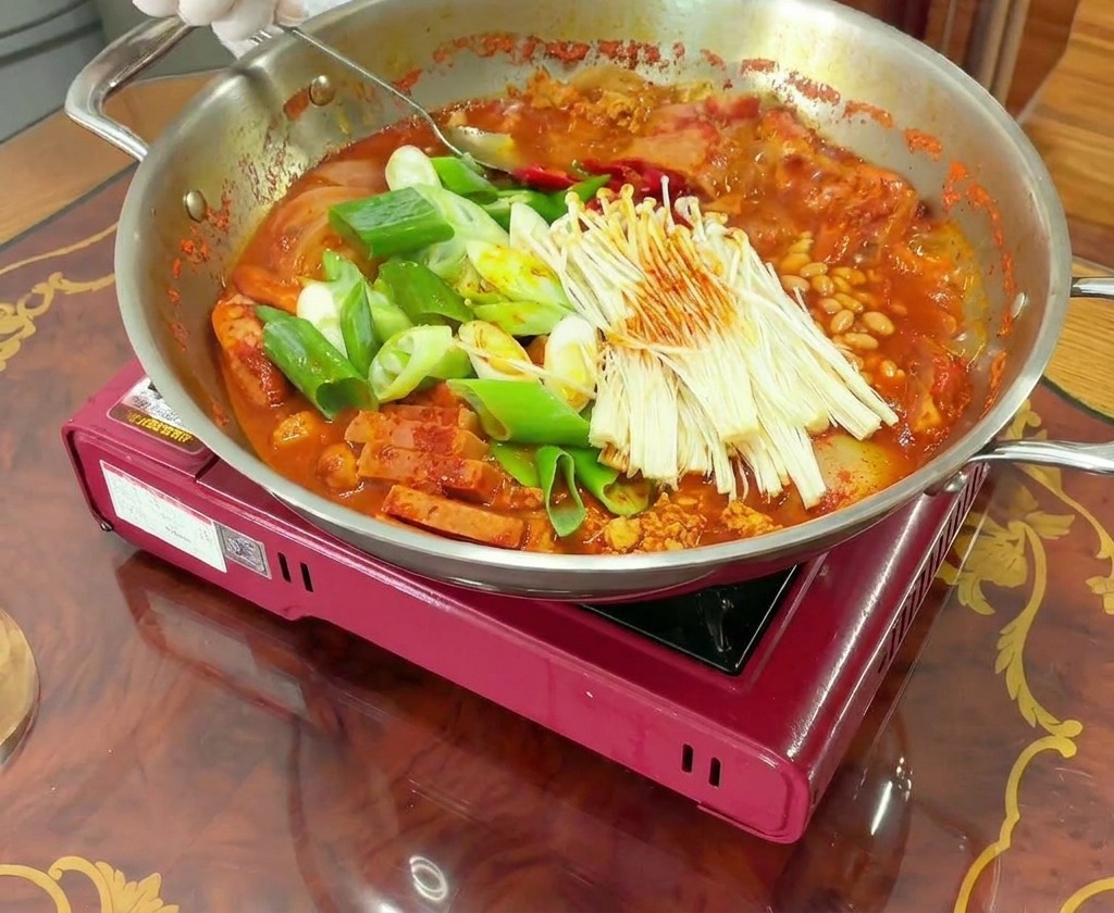 Des mains gantées mélangent avec une cuillère une marmite bouillonnante de Budae Jjigae, liant la viande et les légumes au bouillon rouge.
