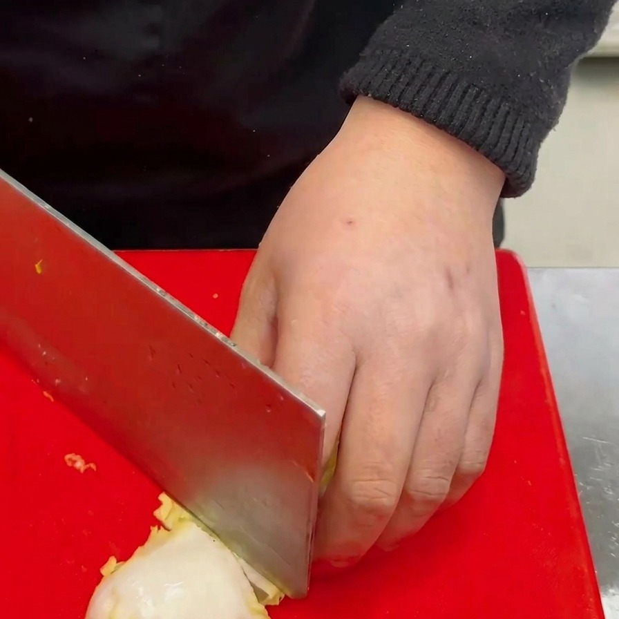 Une main utilisant un grand couperet pour hacher un petit chou blanc en carrés sur une planche à découper rouge.