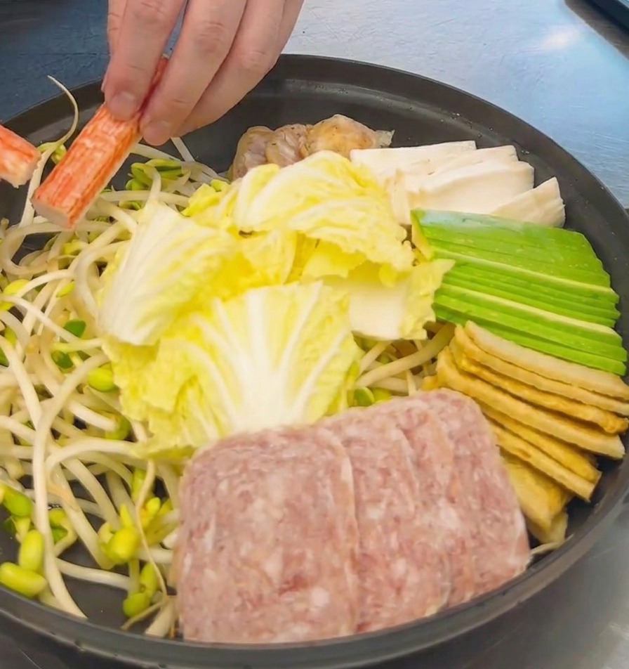 Une main disposant des bâtonnets de crabe orange et blanc dans une marmite remplie de chou, de spam et de tofu.