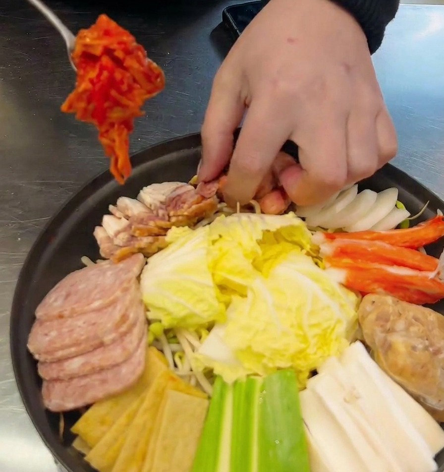 Une cuillère en métal déposant du kimchi rouge dans une marmite garnie de saucisses, de spam et de légumes.