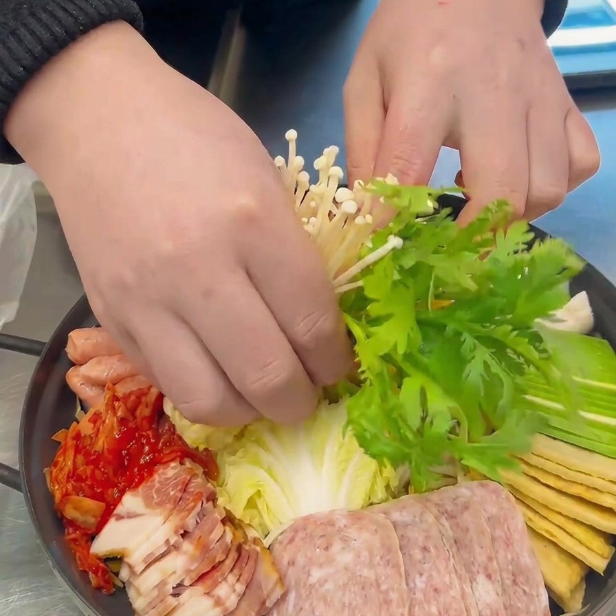 Des mains plaçant des champignons enoki blancs et des feuilles de chrysanthème vertes au centre de la marmite.