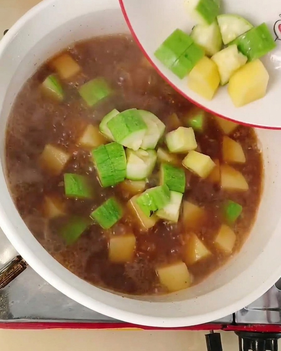 Dés de courgette et oignons versés depuis une assiette dans un bouillon rouge-brun bouillonnant.