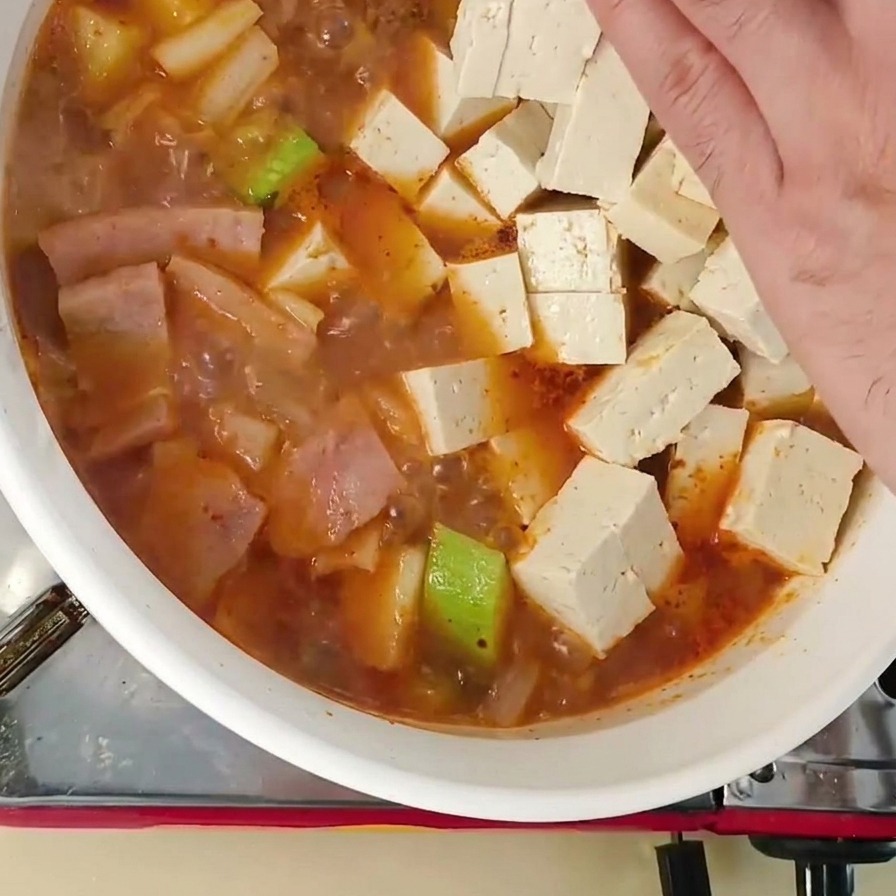 Une main déposant des cubes de tofu frais dans le ragoût coréen contenant courgettes et autres ingrédients.