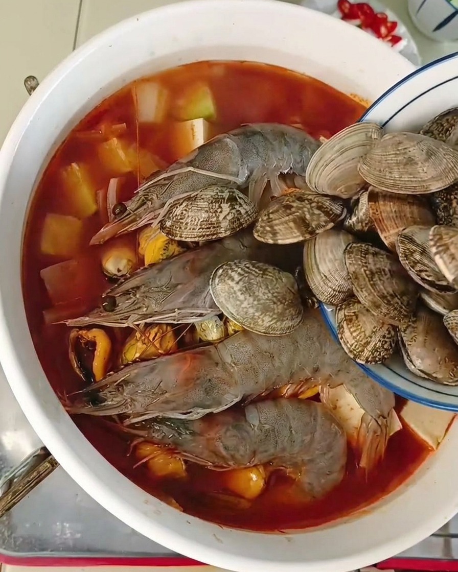 Un bol bleu versant des palourdes fraîches entières dans une marmite remplie de crevettes et de tofu.