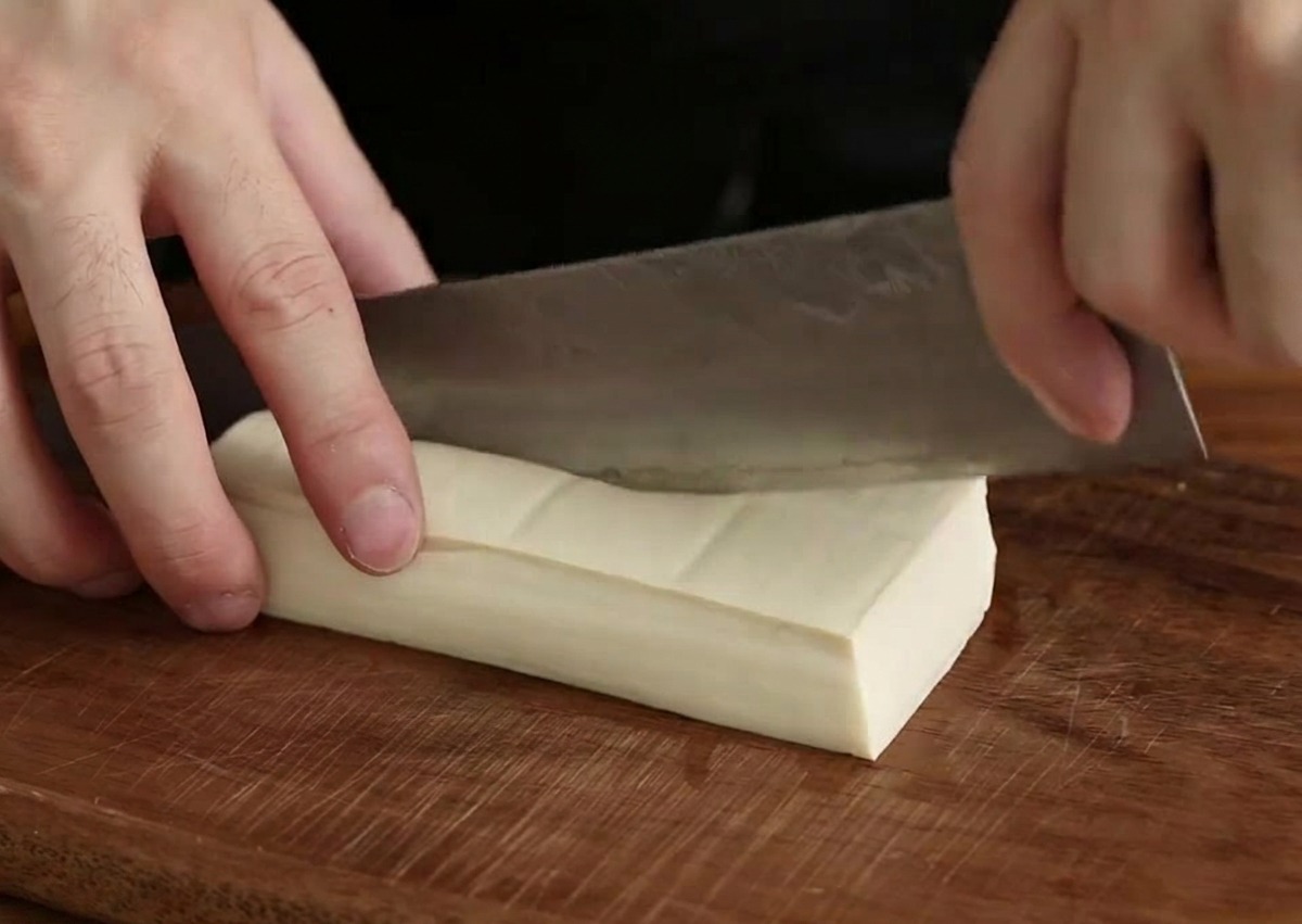 Une main utilisant un couteau de chef pour couper un bloc de tofu blanc en petits cubes sur une planche en bois sombre.