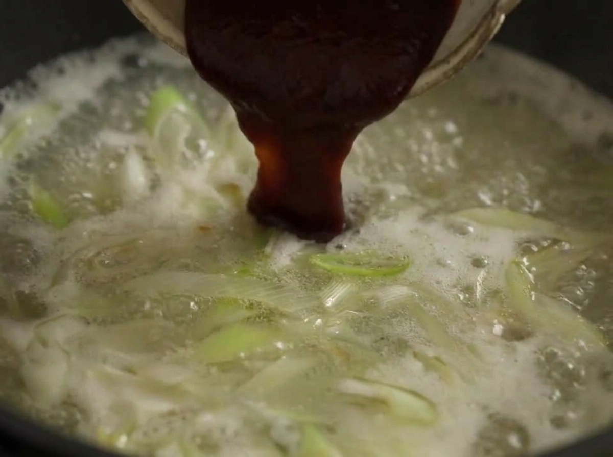 Une grande cuillerée de pâte de piment rouge foncé mélangée dans une marmite de bouillon bouillant avec des poireaux.
