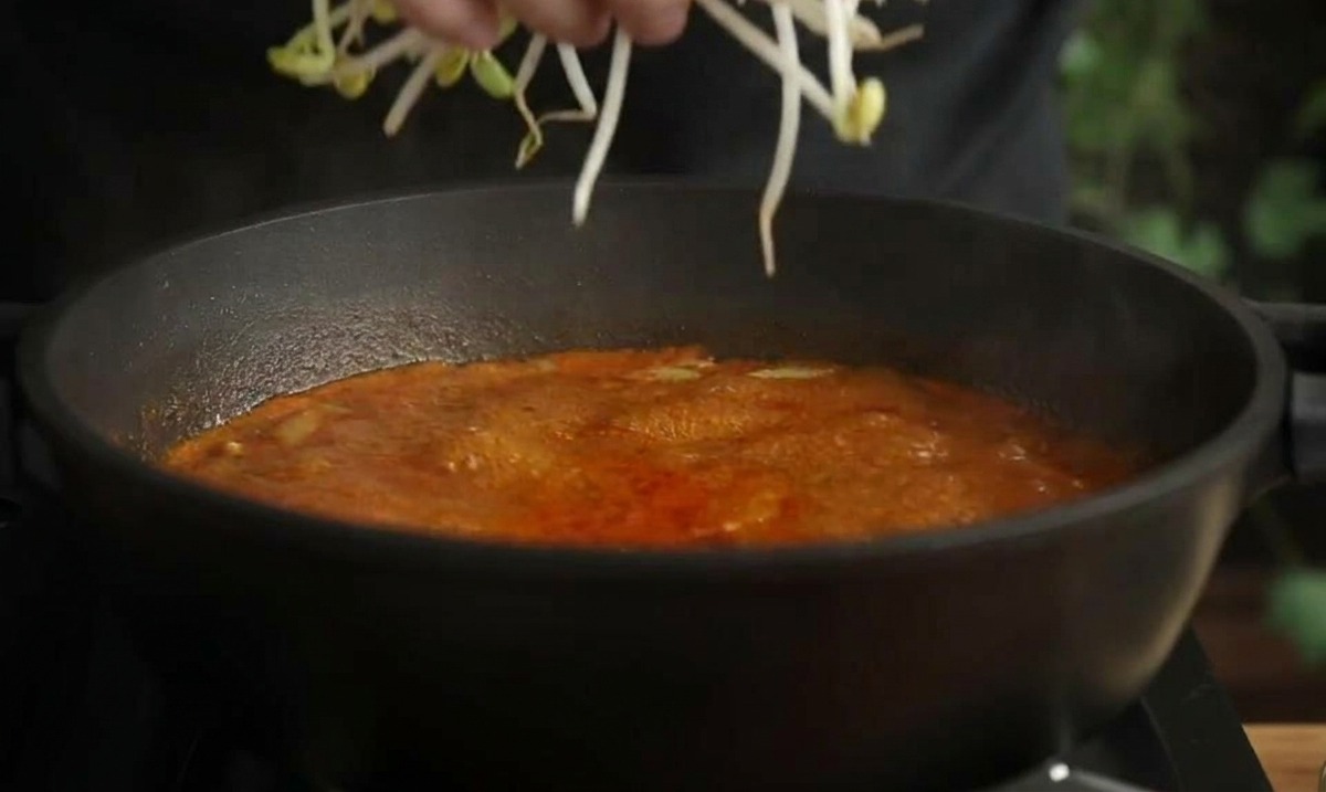 Une main ajoutant une poignée de longues pousses de haricots pâles dans une marmite de soupe de fruits de mer rouge.