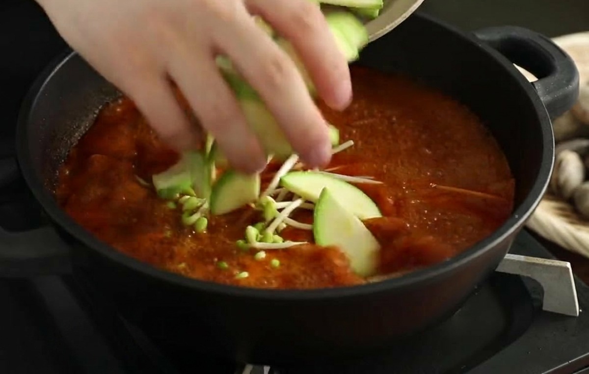 Mains plaçant des tranches de courgette fraîche dans un bouillon de fruits de mer rouge frémissant.