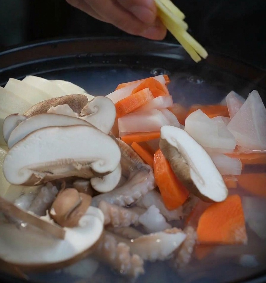 Une main déposant de fines tranches de gingembre frais dans la marmite remplie de légumes et de fruits de mer.