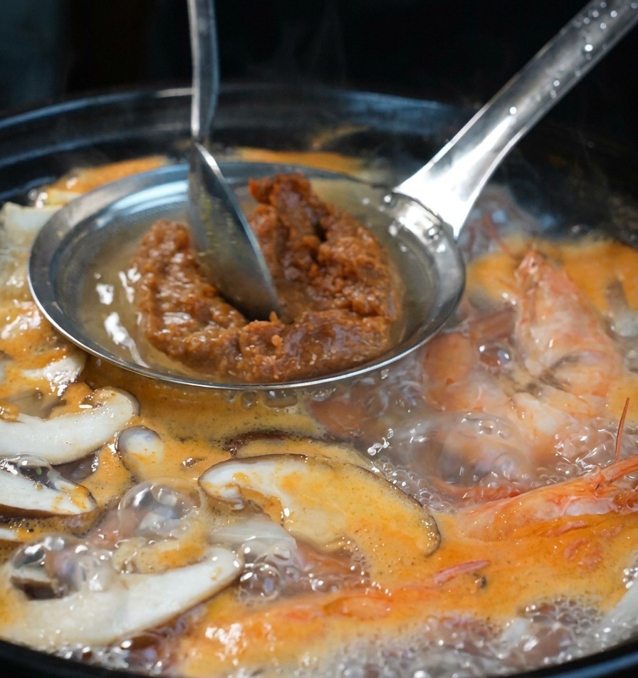 Une main utilisant une cuillère pour presser la pâte de soja à travers un tamis dans le bouillon chaud.