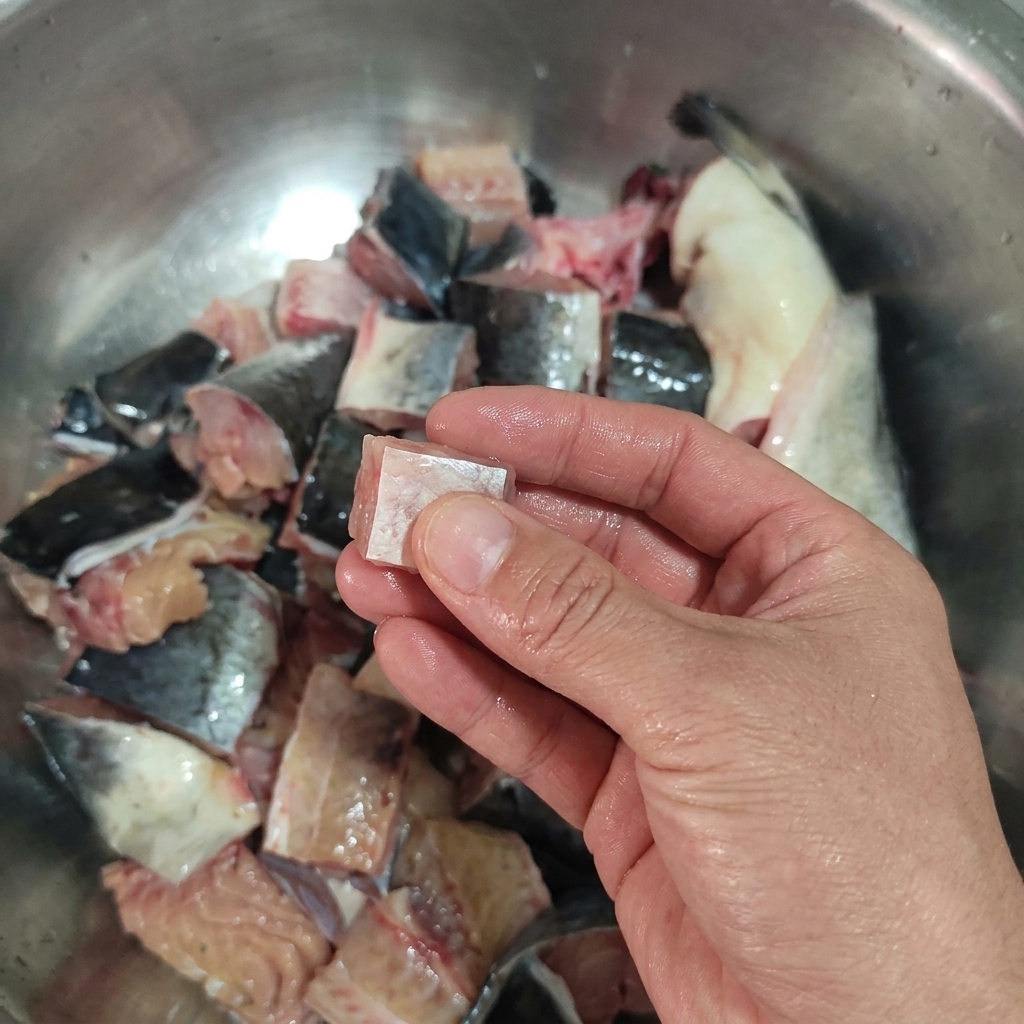 Une main tenant un morceau carré de poisson-chat cru au-dessus dun bol de morceaux déjà coupés.