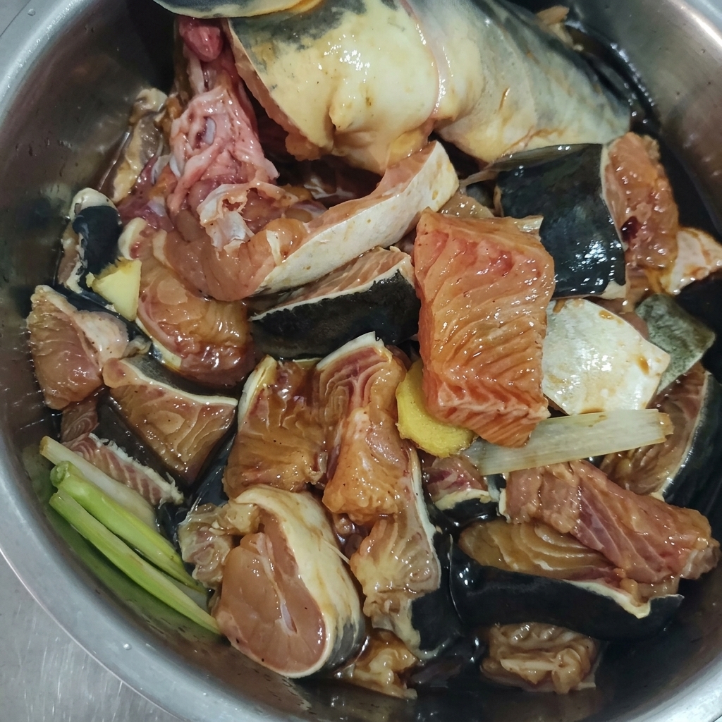 Morceaux de poisson cru mélangés avec du gingembre, des oignons verts et une marinade sombre dans un bol en métal.