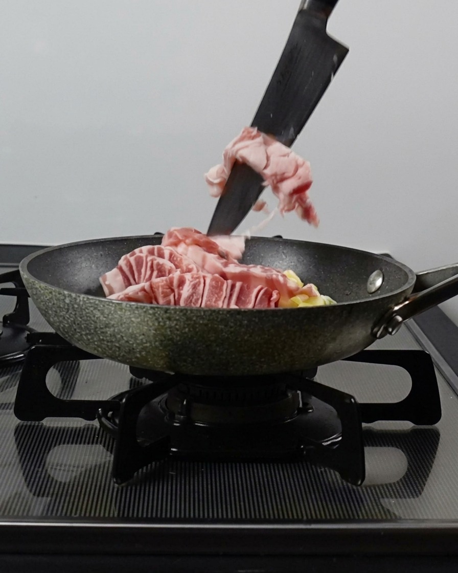 Couteau poussant des lamelles de porc, de lail et des oignons verts dans une poêle chaude.