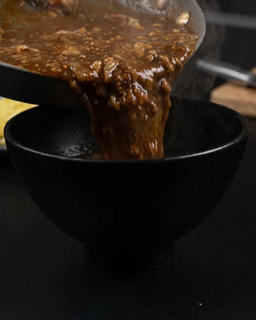 Bouillon épais et sombre versé dune poêle dans un bol noir mat, avec de la vapeur sen échappant.