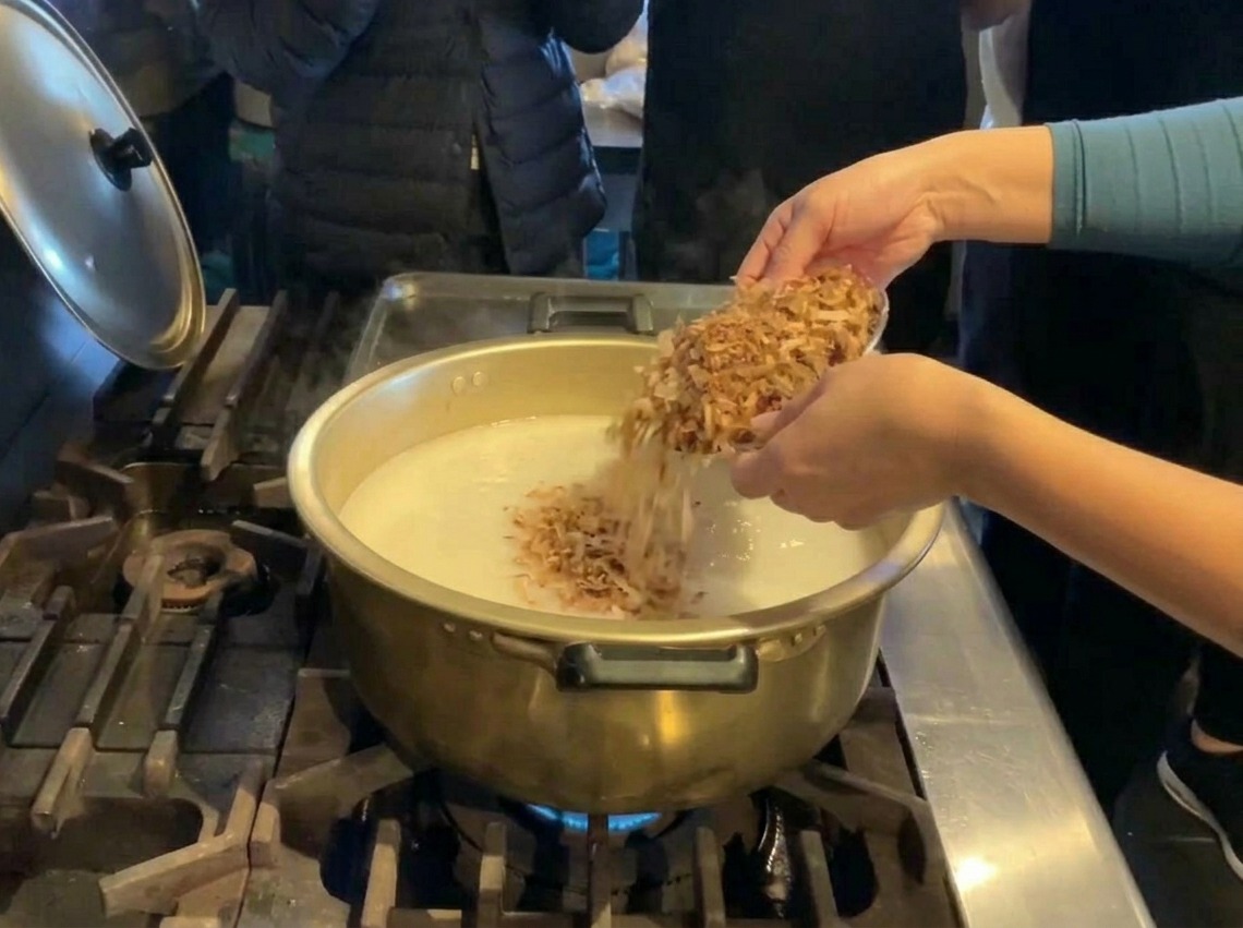 Une personne versant des flocons de bonite séchée dun bol argenté dans une grande marmite de bouillon blanc.