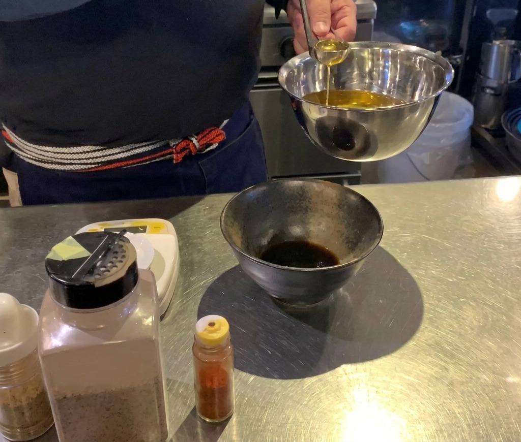 Un cuisinier versant de lhuile dorée avec une petite louche argentée dans un bol noir contenant une sauce sombre.