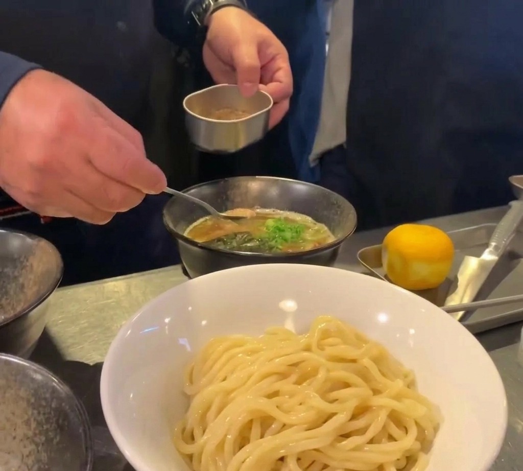Une petite cuillère déposant de la poudre de poisson brune dans un bol de bouillon garni doignons verts.