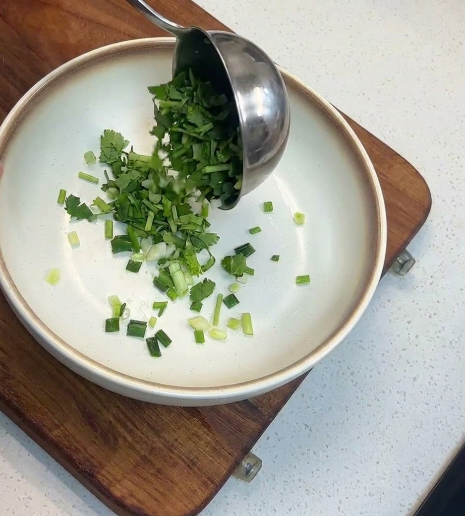 Une louche versant une généreuse quantité de coriandre et doignons verts hachés dans un bol blanc.