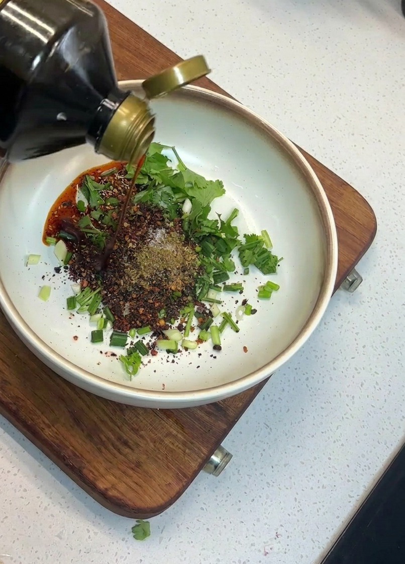 Sauce soja versée dune bouteille en verre dans un bol contenant des herbes et du piment.