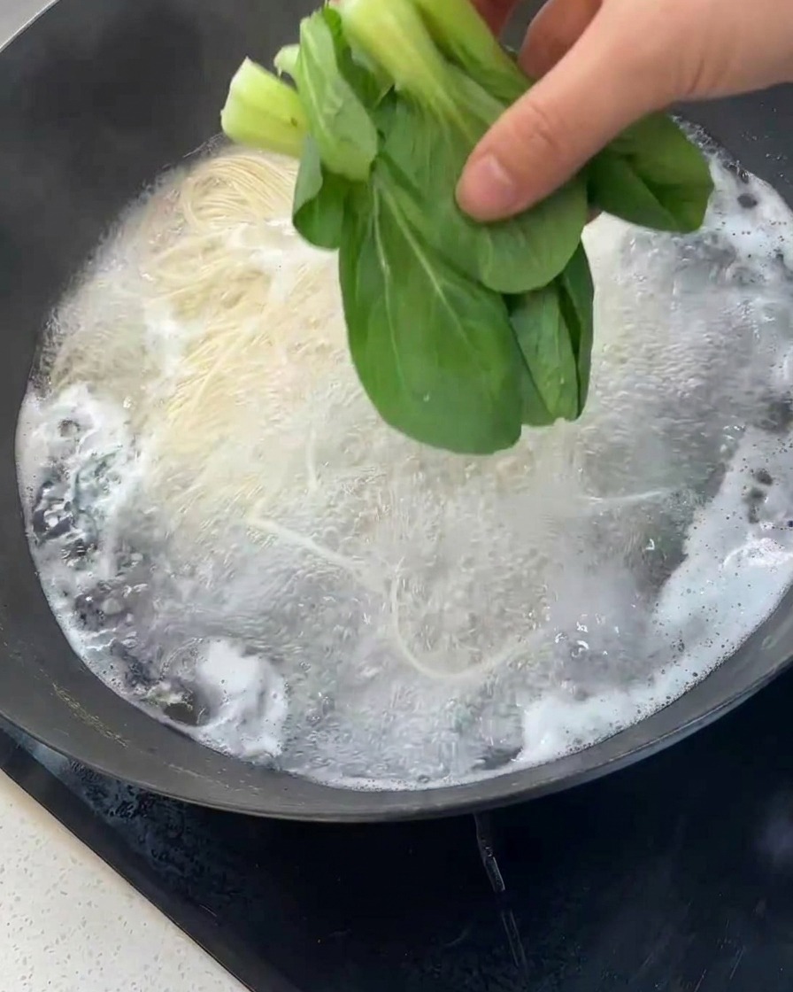 Une main déposant du bok choy frais dans leau bouillante avec les nouilles.