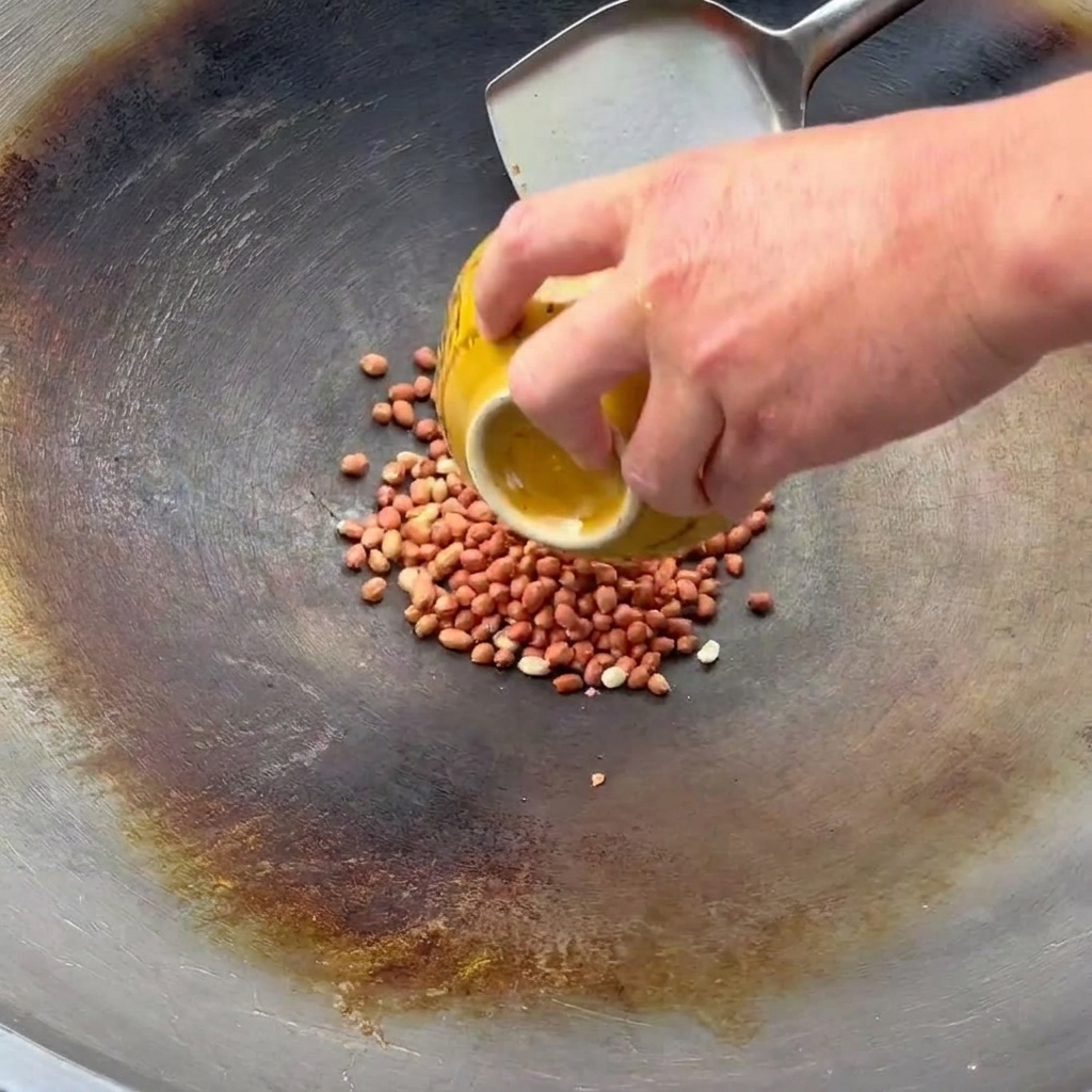 Une main versant des cacahuètes crues dun bol jaune dans un wok pour les faire sauter.
