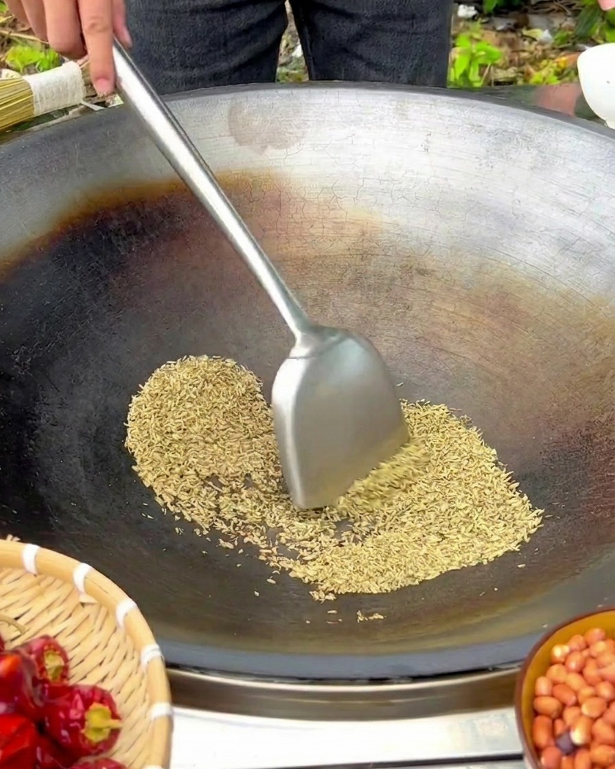 Graines de fenouil remuées avec une spatule dans un grand wok en fonte sur feu doux.