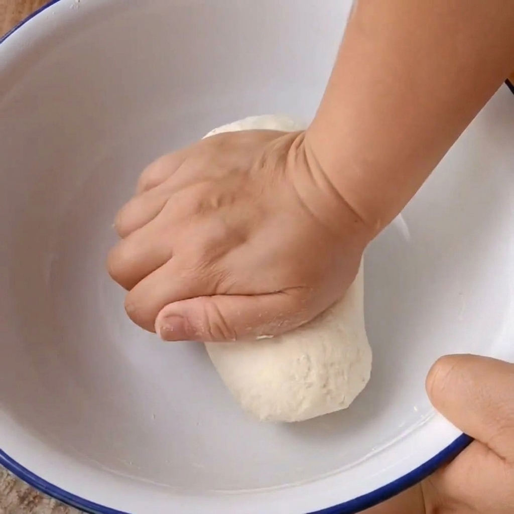 Deux mains pétrissant fermement une boule de pâte dans un bol de mixage blanc.