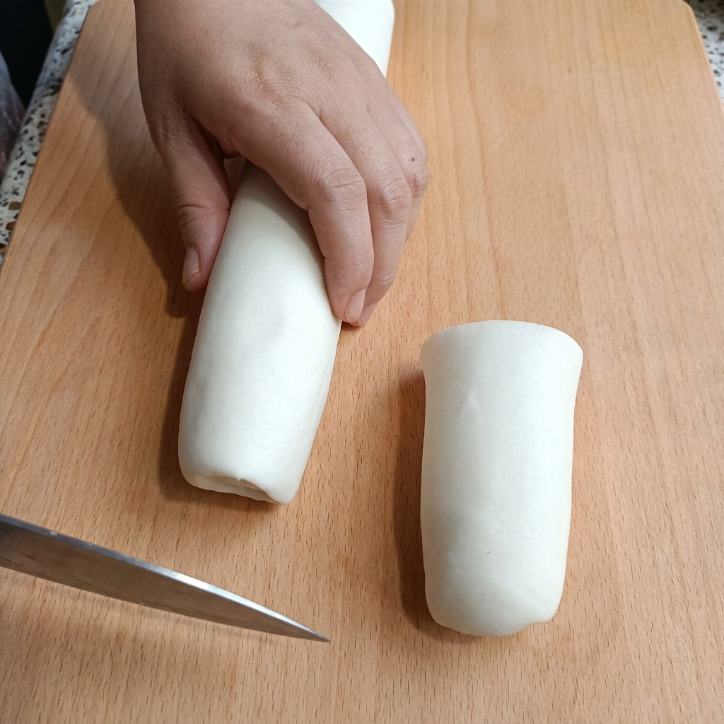 Une main tenant un cylindre de pâte blanche sur une planche à découper en bois tout en le coupant avec un couteau.