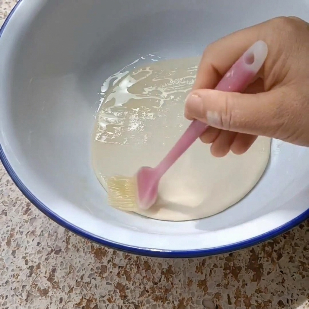 Une main utilisant un pinceau en silicone pour badigeonner un disque de pâte dhuile végétale dans un bol blanc au bord bleu.