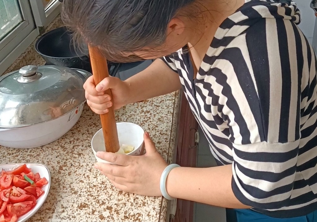 Une personne utilisant un pilon en bois pour écraser des gousses dail en une pâte lisse dans un petit bol blanc sur un plan de travail en granit.