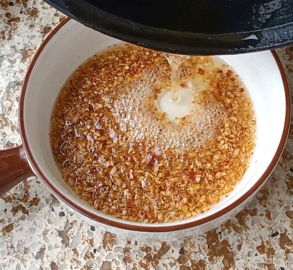 De lhuile chaude versée dune poêle noire dans un bol blanc contenant de lail et des flocons de piment, créant des bulles et de la mousse.