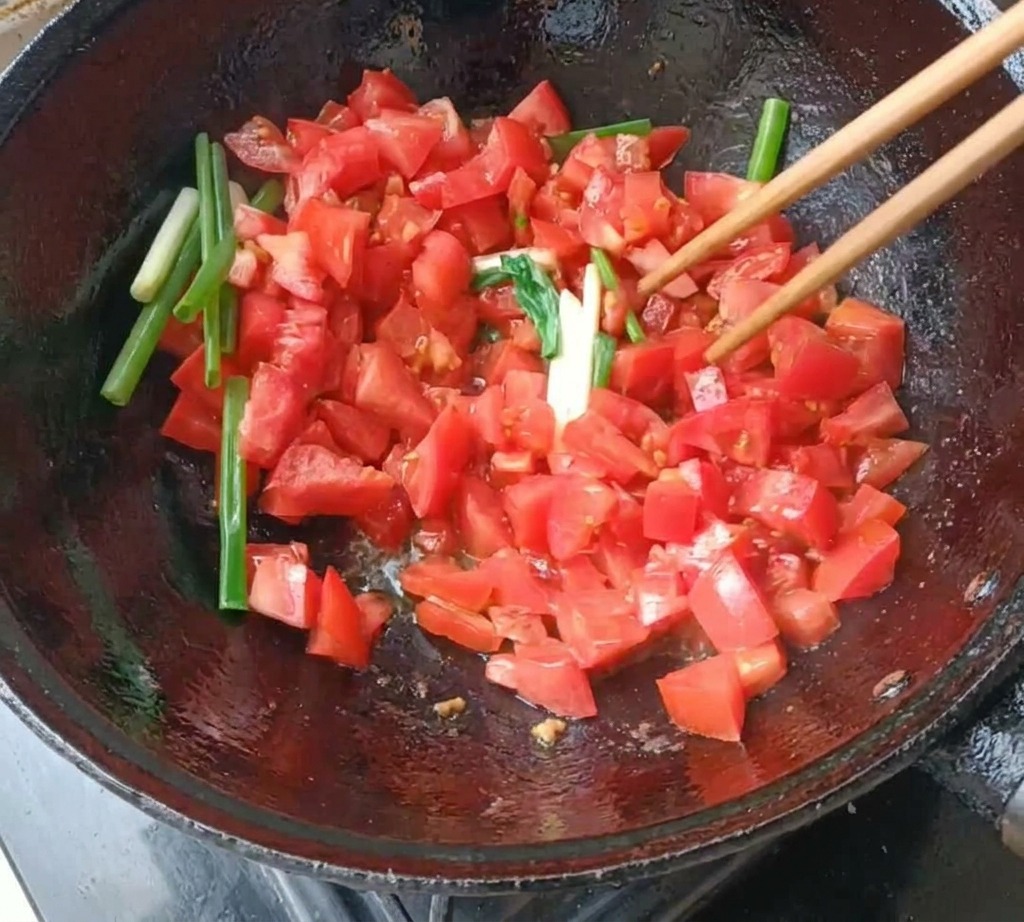 Des tomates en dés et des segments doignons verts sautés avec des baguettes en bois dans un wok en fonte sombre.