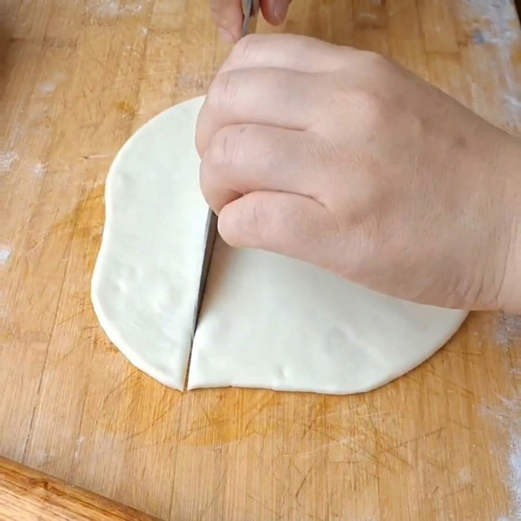 Une main utilisant un grand couteau en métal pour couper un disque de pâte blanc et plat en larges rubans sur une planche à découper en bois.
