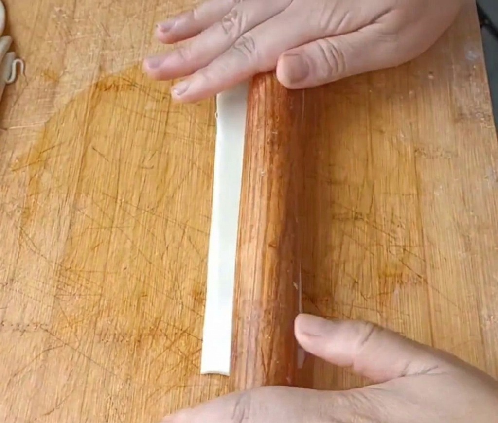 Gros plan sur des mains utilisant un rouleau à pâtisserie en bois pour aplatir une longue lanière de pâte huilée sur une planche à découper.
