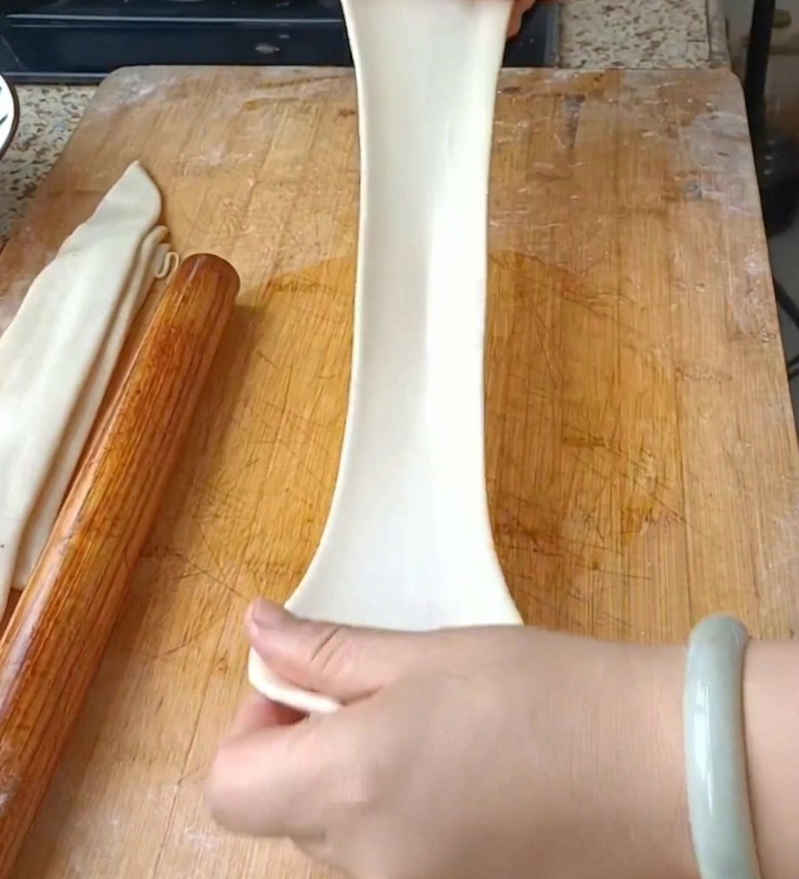 Deux mains tirant verticalement une large lanière de pâte blanche translucide pour former une longue nouille au-dessus dune planche à découper.