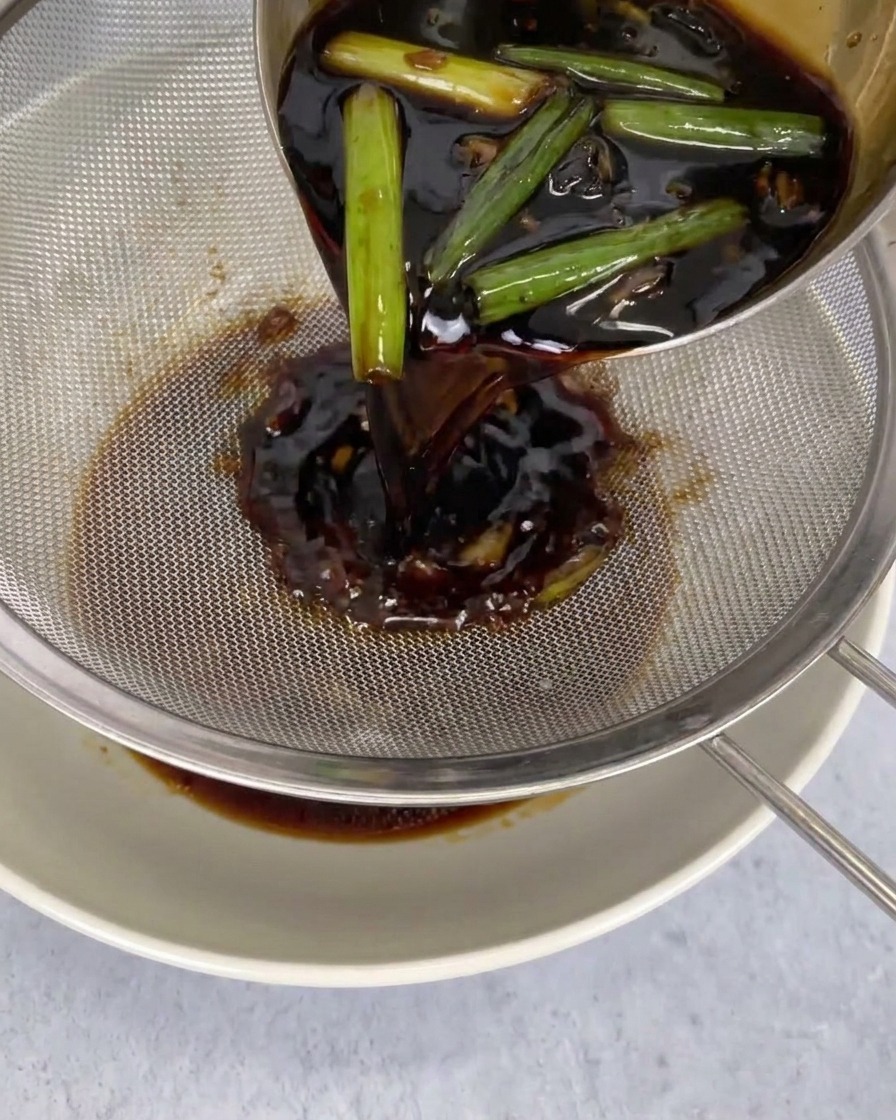 Sauce sombre filtrée à travers un tamis métallique fin dans un bol blanc pour retirer les aromates.
