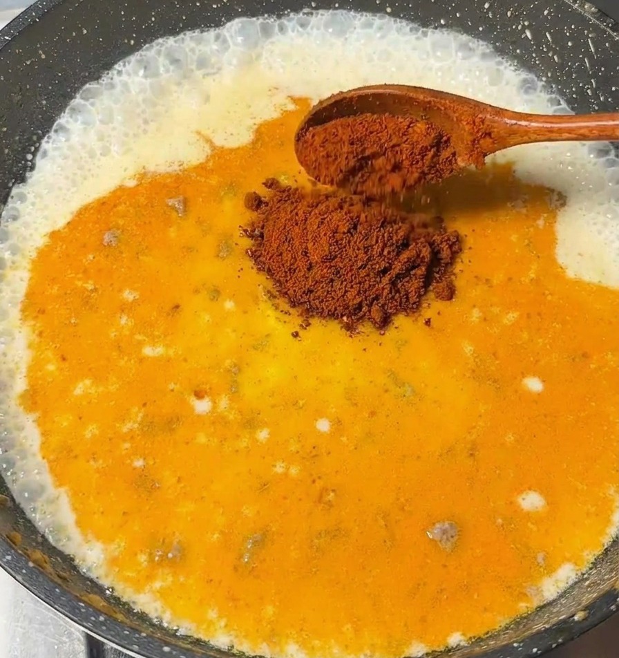 Une cuillère en bois ajoutant des morceaux de roux de curry dans la sauce laiteuse bouillonnante.