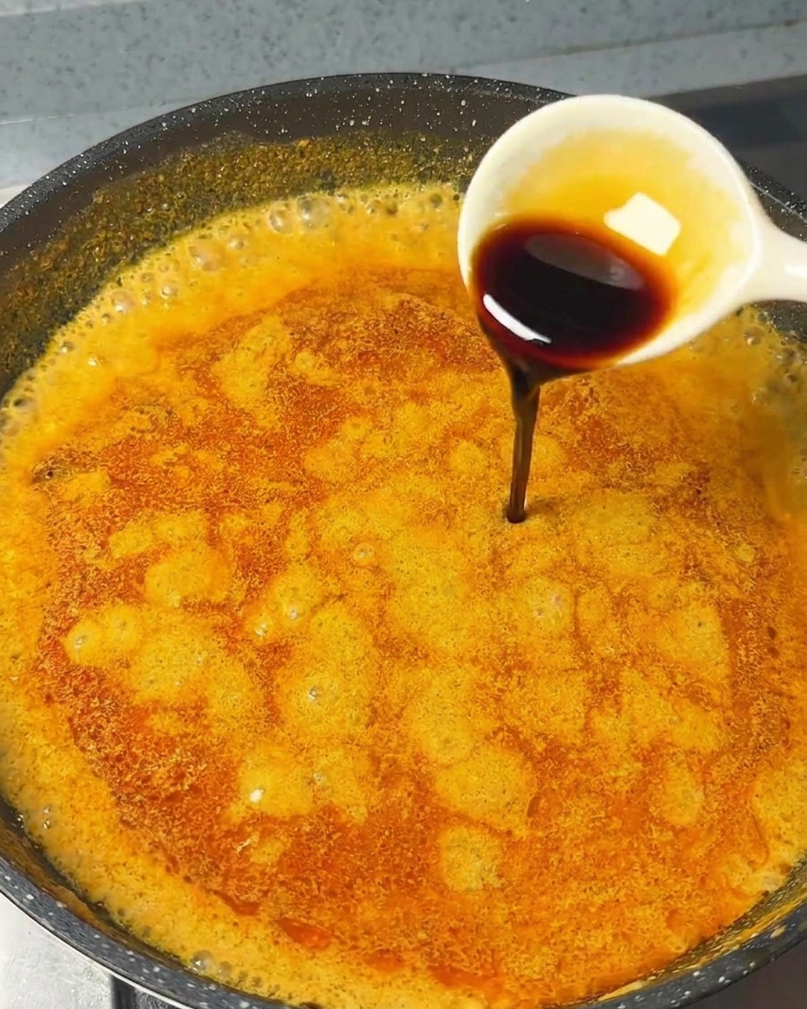 Une cuillère blanche versant de la sauce soja sombre dans la sauce curry orange bouillonnante.