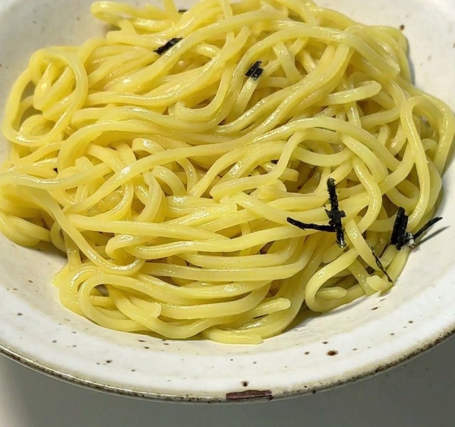 Un bol en céramique blanche rempli de nouilles ramen jaunes surmontées de lanières dalgues séchées noires.