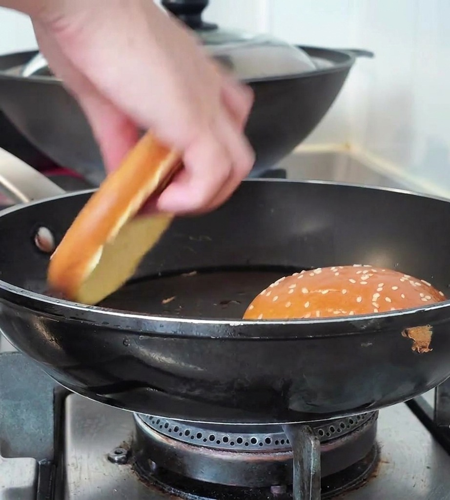 Une main plaçant une moitié de pain au sésame dans une poêle à côté dun autre pain déjà grillé.