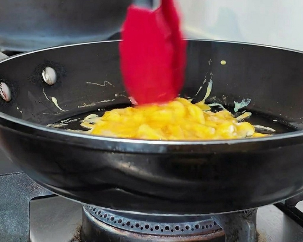 Des pinces rouges rassemblant des œufs brouillés en forme de disque compact dans une poêle.