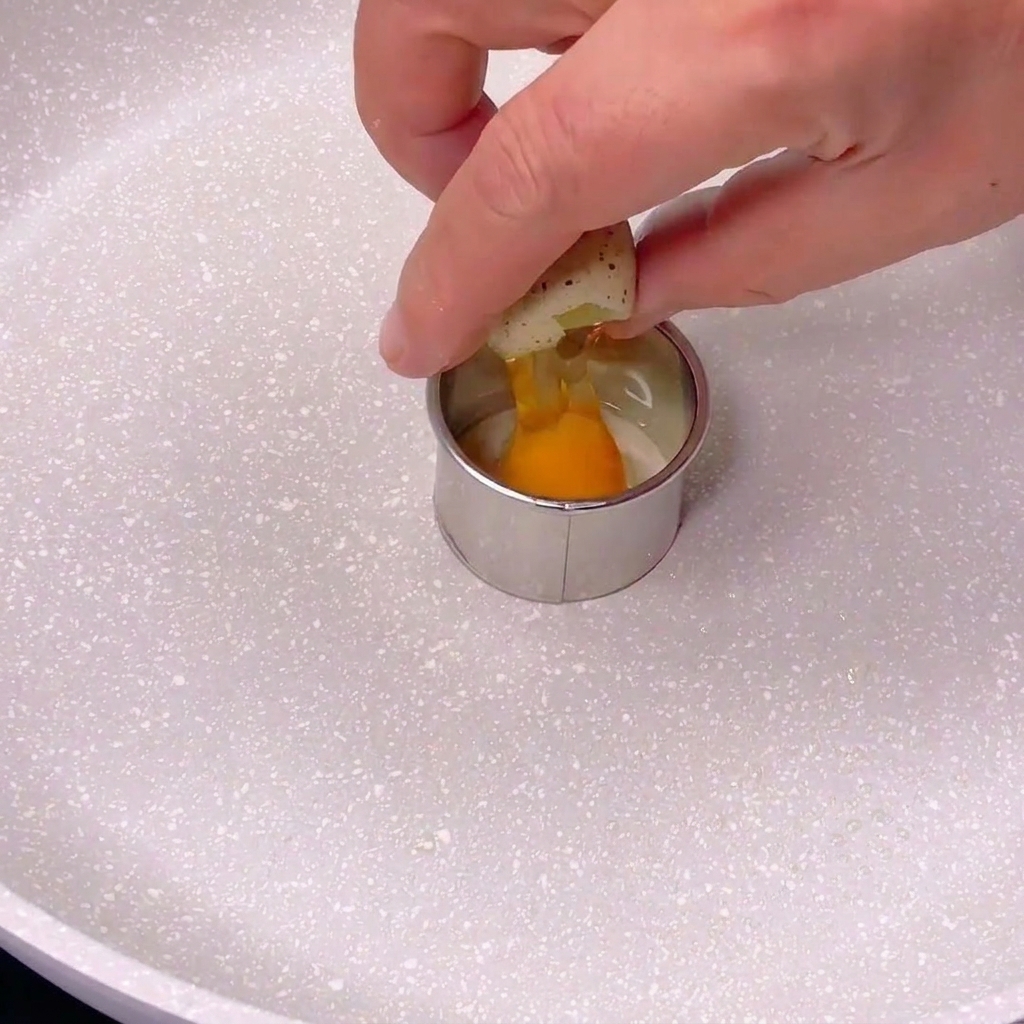 Une personne cassant un petit œuf de caille tacheté dans un moule en métal circulaire posé sur une poêle blanche.