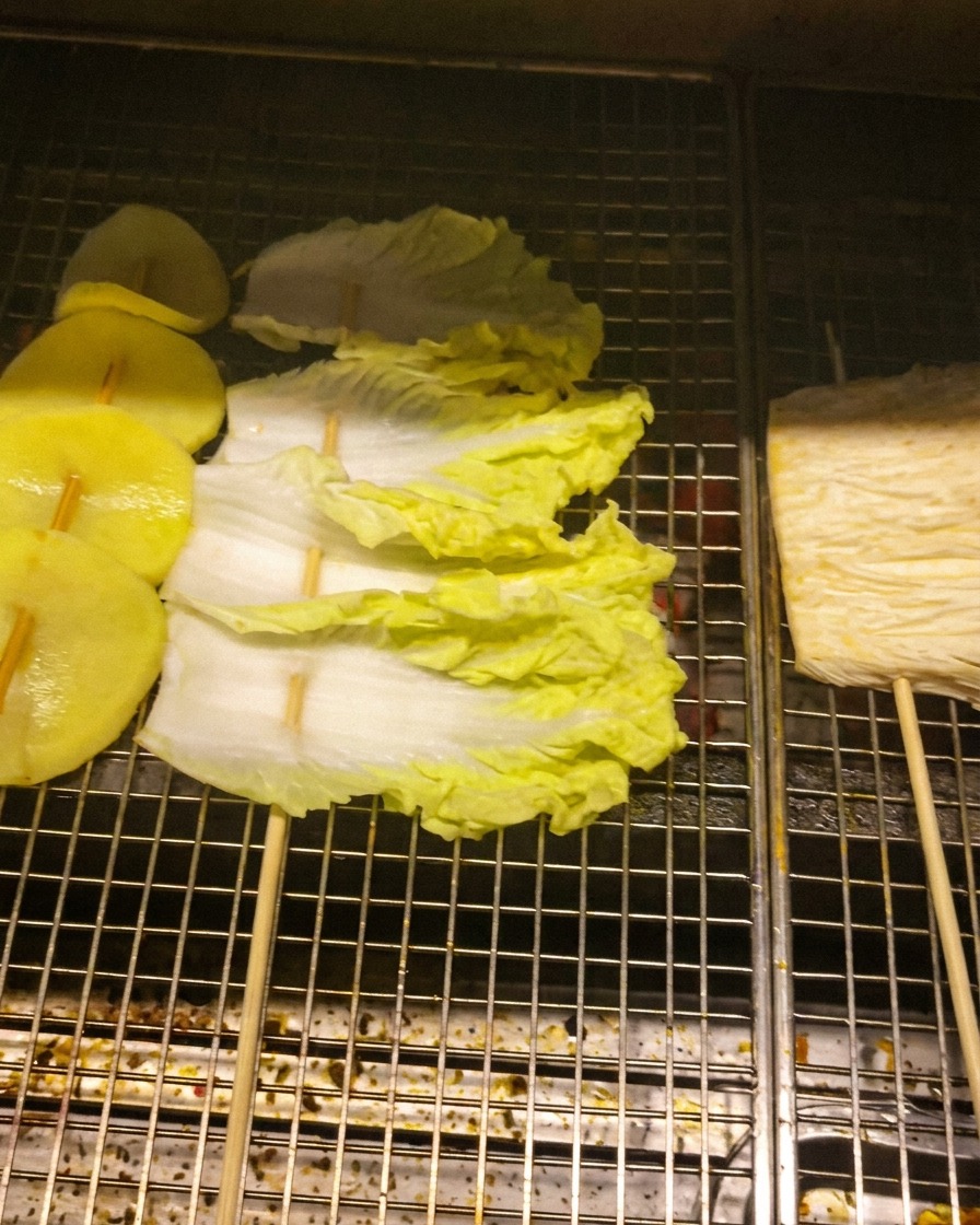 Tranches de pomme de terre, feuilles de chou et champignons enoki enfilés sur des brochettes sur une grille métallique.