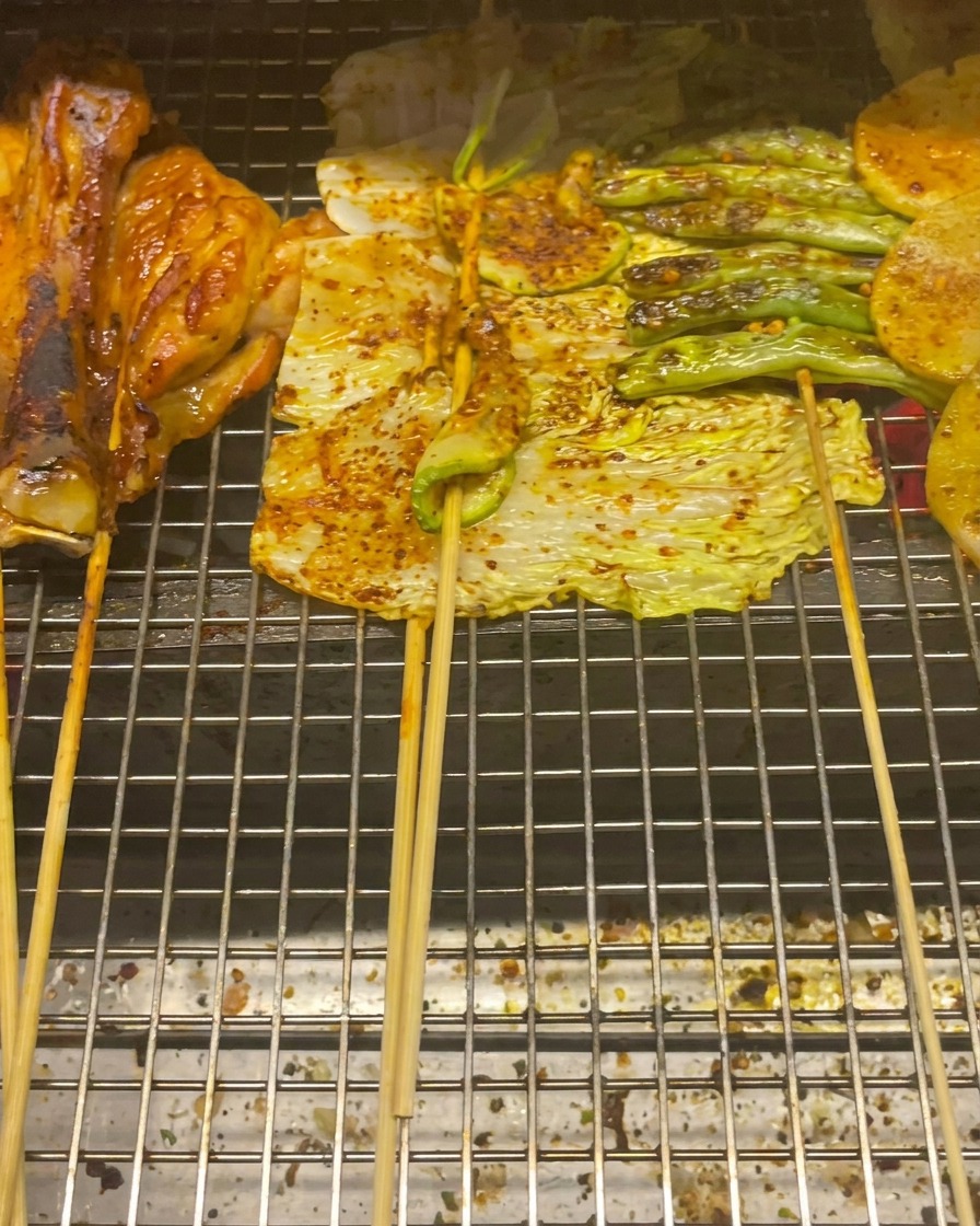 Hauts de cuisse de poulet, petit chou, courgettes, haricots verts et tranches de pommes de terre grillant sur une grille.