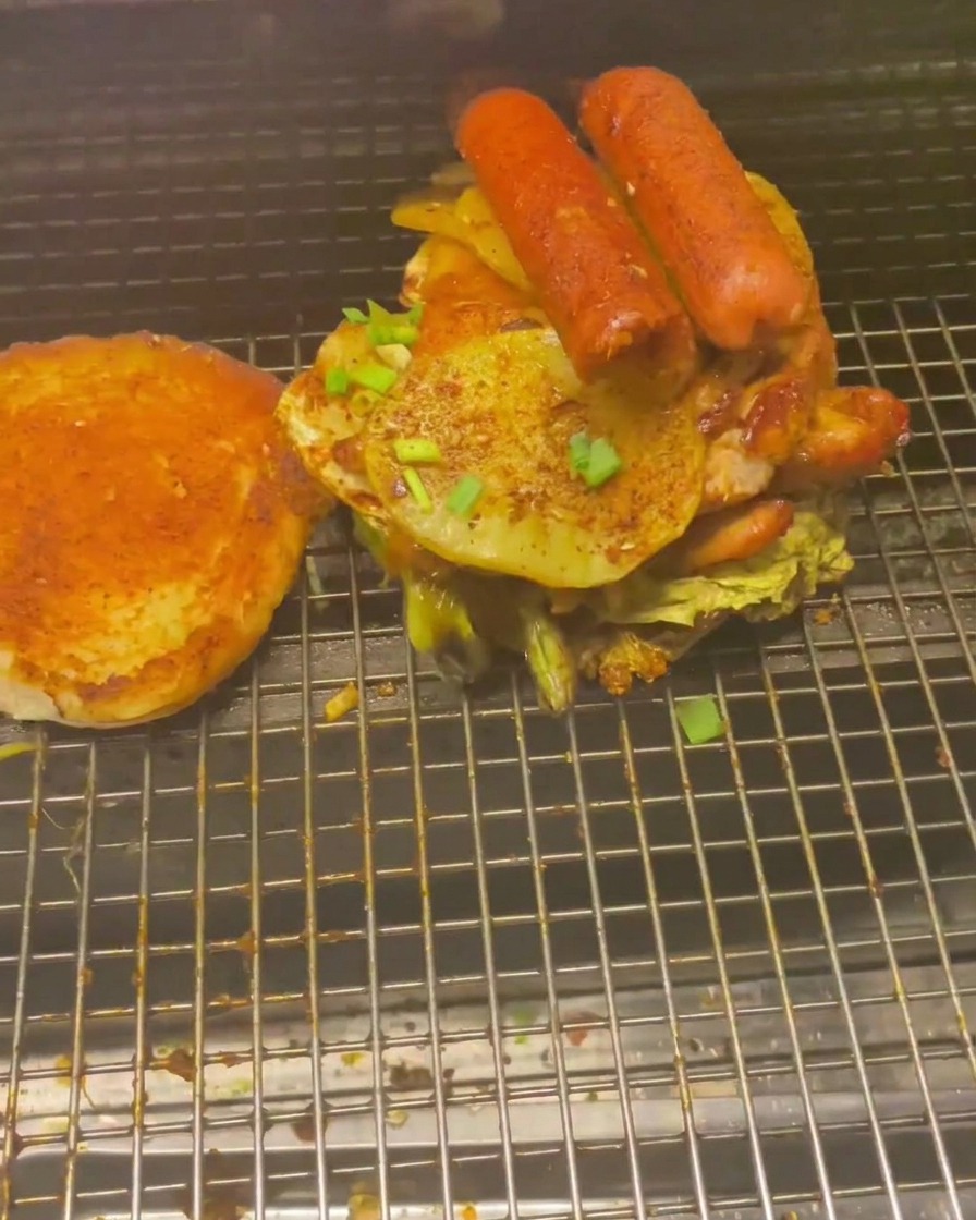Une haute pile de viandes grillées et de légumes, incluant des saucisses et des rondelles de pommes de terre, sur un pain à burger.